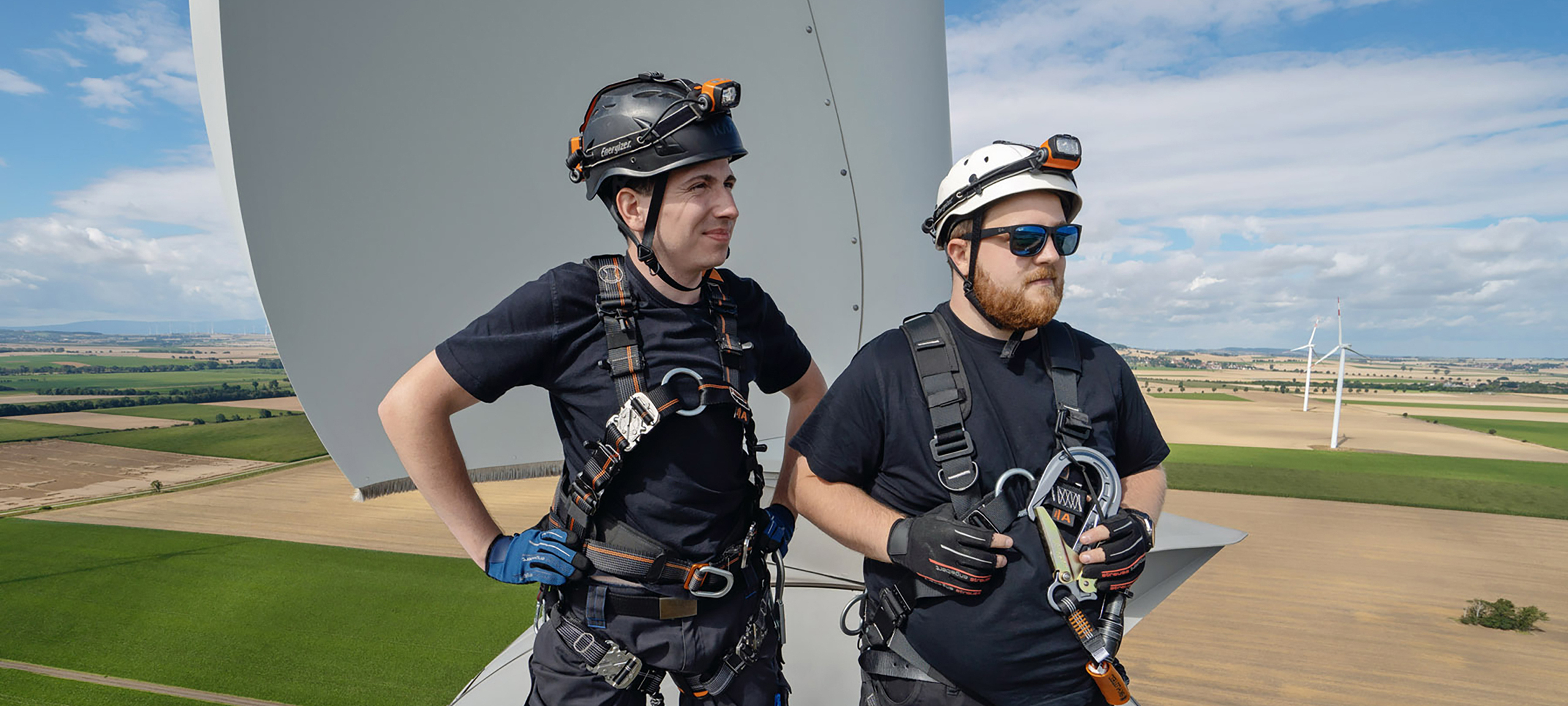 Zwei Mitarbeiter der technischen Betriebsführung stehen auf dem Maschinenhaus einer Windenergieanlage. Sie tragen eine persönliche Schutzausrüstung und blicken nach rechts.